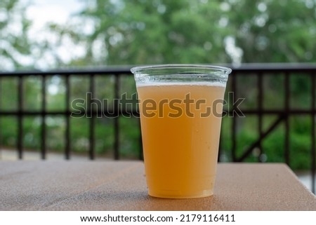 A tall clear plastic glass of sour craft beer in a microbrewery. The cold refreshment has frost on the glass. The beer is on a wooden table. The top of the glass has white froth around the rim. 