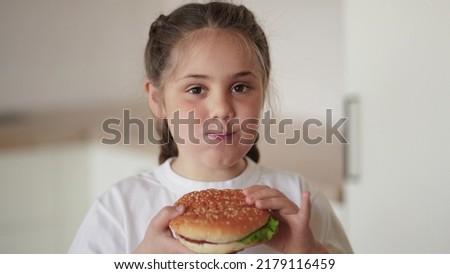 little girl eating a hamburger. unhealthy fast food proper nutrition concept. child greedily with pleasure bites a big burger in the kitchen at home lifestyle. kid eats fast food close-up