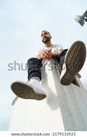 full body of young male model with beard and short hair sitting relaxed wearing white t-shirt, jeans and sport shoes, casual urban fashion on sky background, attractive latin man