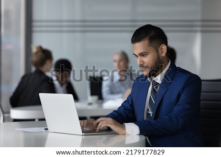 Focused Indian employee man working on online project at laptop, typing, chatting, sitting at workplace table in open office with business meeting of diverse colleagues in background Royalty-Free Stock Photo #2179177829