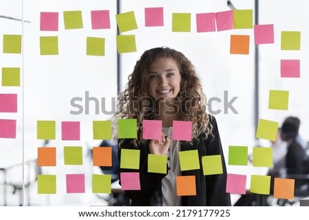 Happy young female business leader head shot through glass portrait. Cheerful young pretty intern girl, employee working in project at paper notes on glass board, looking at camera, smiling