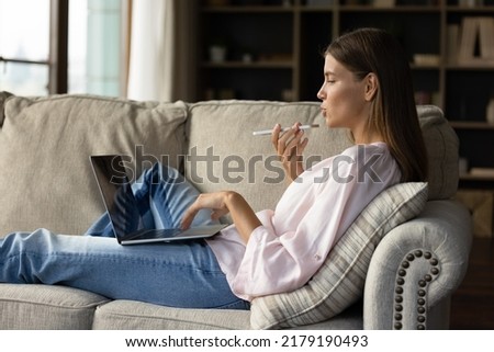 Side view woman holding cellphone makes personal or business call talks on speaker phone, web surfing information using laptop sit on sofa at home. Remote communication, young gen and AI tech concept