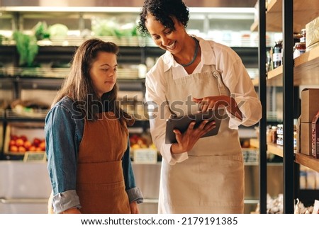 Happy grocery store manager using a digital tablet while having a discussion with her employee. Female shop owner giving a woman with Down syndrome training in managing online grocery orders. Royalty-Free Stock Photo #2179191385