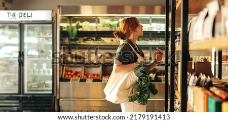 Female shopper choosing food products from a shelf while carrying a bag with vegetables in a grocery store. Young woman doing some grocery shopping in a trendy supermarket.