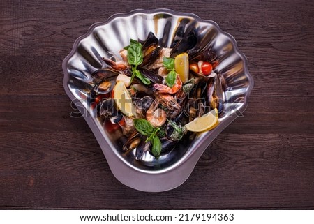Seafood salad of grilled mussels, shrimps and squid. On top are basil leaves and cherry tomatoes. The food is in a metal shell-shaped form. The shape stands on a wooden background.