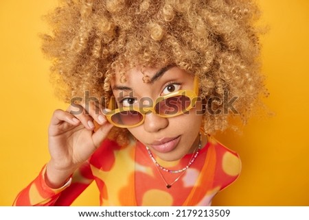 Headshot of serious woman with curly hair looks from sunglasses dressed in long sleeved jumper isolated over vivid yellow background listens attentively information. People and style concept