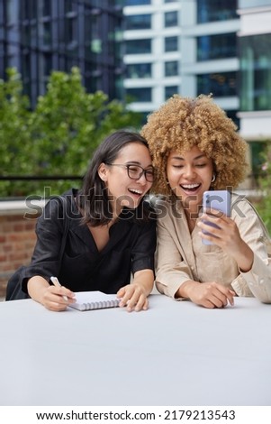 Cheerful female friends make selfie on smartphone for sharing in social networks prepare for exams together make notes in spiral notebook sit at table against urban background. Meeting together