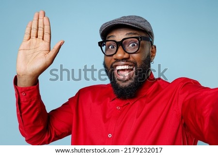 Close up of excited young bearded african american man in red shirt eyeglasses cap doing selfie shot on mobile phone waving greeting with hand isolated on pastel blue color background studio portrait