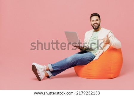 Full body young smiling man in trendy jacket shirt sit in bag chair hold use work on laptop pc computer show thumb up isolated on plain pastel light pink background studio. People lifestyle concept