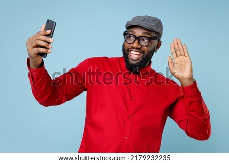 Funny young bearded african american man 20s in casual red shirt eyeglasses cap waving greeting with hand doing selfie shot on mobile phone isolated on pastel blue color background studio portrait