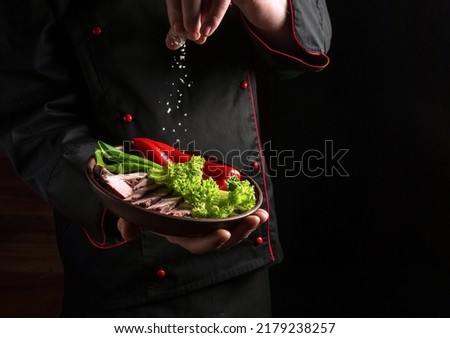 The chef sprinkles salt on the meat and vegetables on the plate. Food preparation concept for hotel on dark background