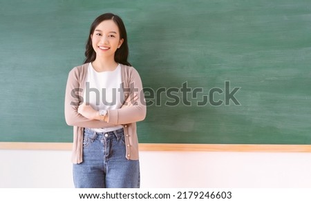 Portrait of smiling beautiful Asian teacher standing with arms crossed looking at camera in front of a blank chalkboard in classroom. Back to school concept, with copy space Royalty-Free Stock Photo #2179246603