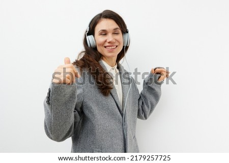 funny funny woman in stylish coat and headphones enjoying music gesturing signs of approval while standing on a light background with empty space