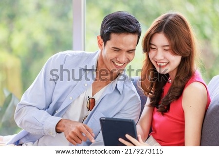 Asian young happy lovely boyfriend and girlfriend couple in casual outfit sitting smiling on cozy sofa couch in living room using tablet computer watching streaming movie online together in weekend.