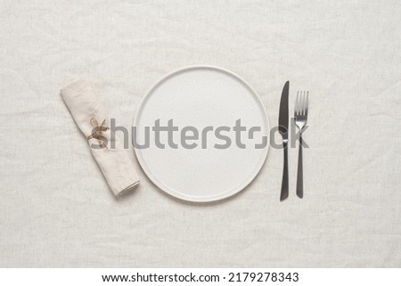 Table setting. Empty beige plate, cutlery and beige linen napkin on beige linen tablecloth. Top view, flat lay. Textured object, selective focus. Royalty-Free Stock Photo #2179278343
