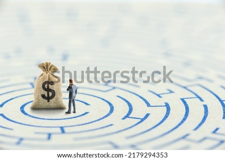 Reducing or overcome financial barrier, financial concept : US dollar bag on a maze puzzle with a businessman considers how to overcome financial difficulties. CFO wishes to avoid a financial crisis. Royalty-Free Stock Photo #2179294353