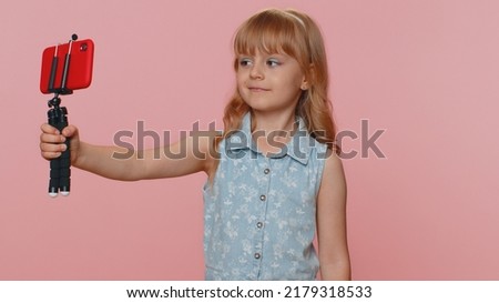 Young preteen child girl kid blogger taking selfie on smartphone selfie stick, communicating video call talking online with subscribers. Little toddler children isolated on studio pink background