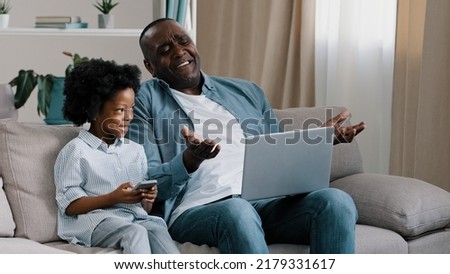 Upset male freelancer sitting on sofa in room with laptop dissatisfied with result loses online game gets bad news little cute daughter sitting next to dad smiling holding mobile phone watching father