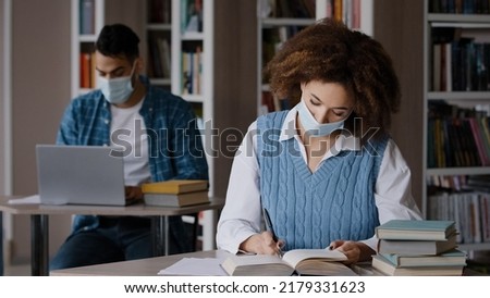 Students study in library in protective mask comply with quarantine measures young clever girl student reading book looking for information in textbook writes notes preparing for exam doing homework