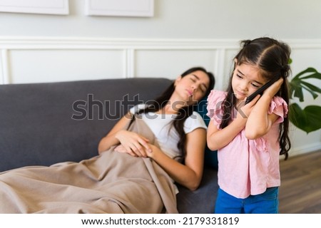 Worried little girl calling a doctor on the phone and asking for help because her sick mother is in pain lying on the sofa at home 