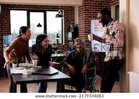Diverse team of smiling workers and paralyzed man taking break from work in business office. Male employee wheelchair user suffering from physical health condition laughing with colleagues. Royalty-Free Stock Photo #2179334083