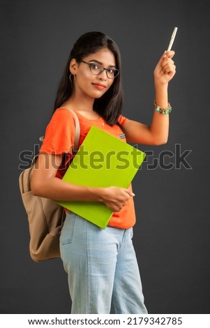 Beautiful young girl with a backpack standing and holding a notebook, posing on a grey background.
