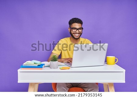 Happy African man using laptop while sitting at the desk against purple background