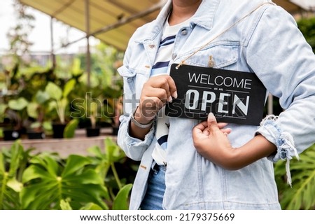 Close up, Gardener farmer standing in ornamental plants shop with text “welcome we are open”. Happy woman owner showing open sign in her small business shop. Your welcome to my small business