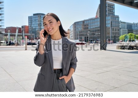 Young Asian successful businesswoman leader wearing suit standing in big city talking on mobile phone. Smiling woman making business call on cell walking on busy downtown street outdoors. Royalty-Free Stock Photo #2179376983