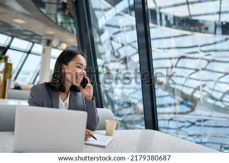 Young happy Asian business woman company employee or sales manager wearing suit working in contemporary glass office talking to client on the phone using laptop working in big modern work space. Royalty-Free Stock Photo #2179380687