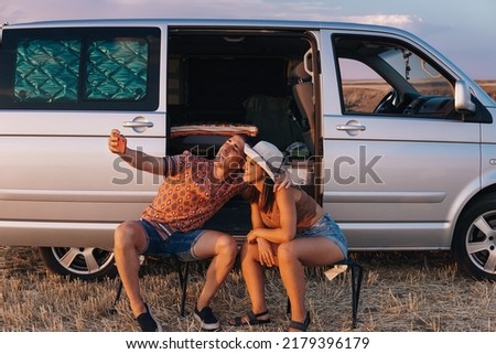 Young couple consisting of boy and girl sitting in camping chairs outside their camper van. Both are trying to take a selfie while smiling and putting their heads together. She is wearing a white hat.