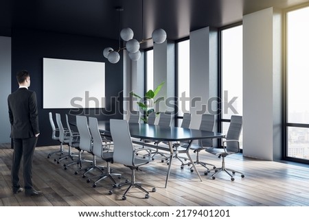 Attractive young european businessman standing in modern meeting room interior with panoramic city view and empty white mock up banner. Worker and CEO concept