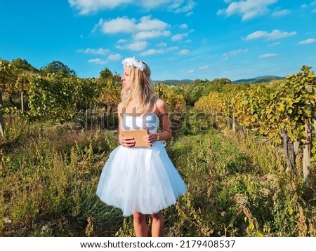 young woman standing in vineyard and looking around. girl wearing white wedding dress and flower wreath on head. lady holding book or diary. nature autumn wallpaper. beautiful bride.