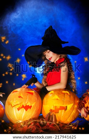 Little girl in a costume of witch posing with pumpkins over dark background.
