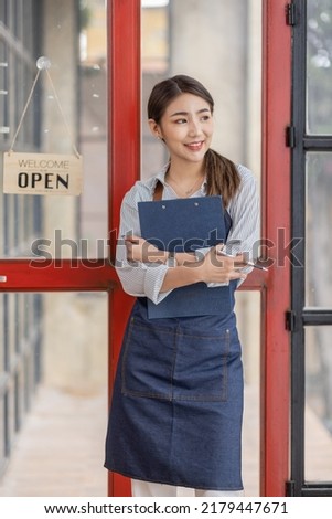 Shot of smiling young cafe show owner asian woman standing with arms crossed in the doorway. Open sign on the glass door.