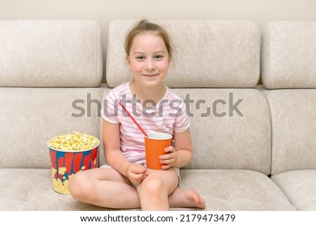 joyful cute little girl sitting on the couch at home with a big glass of popcorn and a glass of drink in her hand