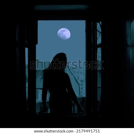 full moon in the night dark  blue  sky. Black silhouette of young slim magical whitch woman looking out of an open balcony window. city, town with many houses on background. Royalty-Free Stock Photo #2179491751