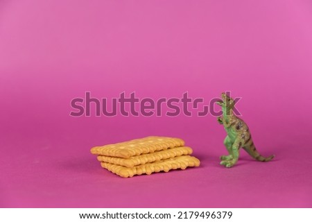 Dinosaur and crackers against a purple background. Rectangular crispy crackers. Green miniature of a predatory animal standing on its hind legs. Selective focus.