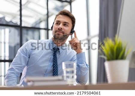 Cheerful young support phone male operator in headset, at workplace with collegues on the background while using computer, help service and client consulting call center concept