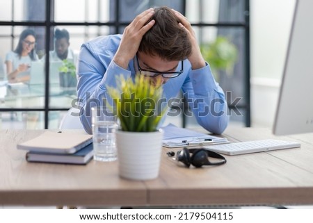 Unhappy frustrated young male holding head by hands sitting with computer behind desk at office with collegues on the background