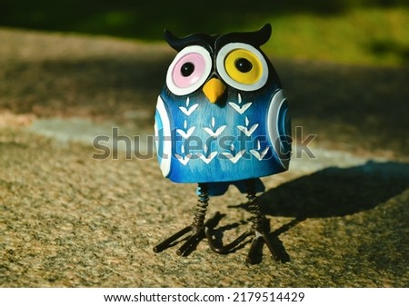 A close-up toy of a surprised blue owl with multi-colored eyes stands on a granite stone. The toy is illuminated by evening sunlight and casts a shadow