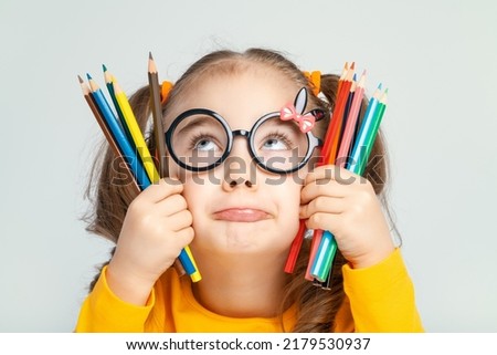 Beautiful cute little preschooler girl with glasses holding colorful pencils and pouting her lips. Sulky child with pencils. Imagination and creativity at school concept.  Royalty-Free Stock Photo #2179530937