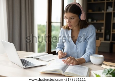 Diligent student woman studying sit at desk with laptop and textbook, makes notes, writing essay looks concentrated. Self-education, e-learning, university exams or admission preparation, tech concept Royalty-Free Stock Photo #2179531111