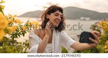 Cheerful young caucasian woman in good mood takes pictures of herself on phone on backdrop of landscape at sunset. Brunette wears casual clothes in summer. Relaxation concept, technology
