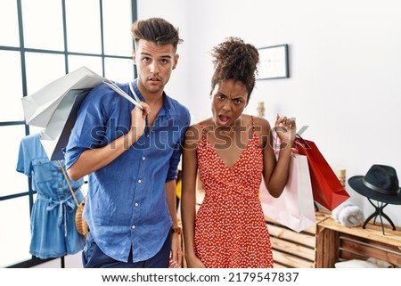 Young interracial couple holding shopping bags at retail shop in shock face, looking skeptical and sarcastic, surprised with open mouth 