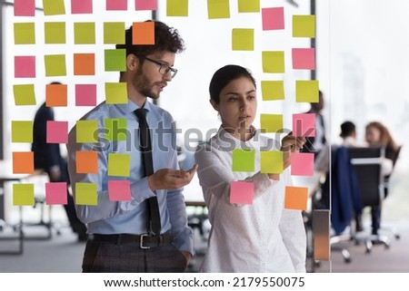 Diverse millennial employees, coworkers discussing project schedule, writing memo on sticky notes. Startup leader, coach, training young Intern guy, using Kanban board for planning tasks Royalty-Free Stock Photo #2179550075