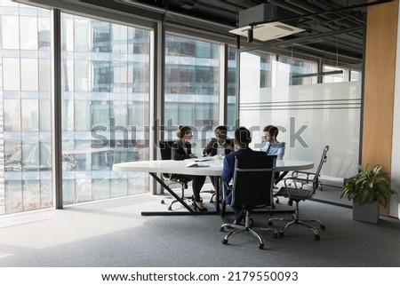 Diverse millennial business team talking in meeting room, negotiating on project at table at glass wall panoramic window, discussing deal in open space, modern office interior. Wide shot Royalty-Free Stock Photo #2179550093