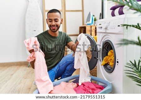Young hispanic man cleaning clothes using washing machine at laundry Royalty-Free Stock Photo #2179551315