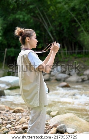 Attractive young Asian female taking a picture of the beautiful forest and river with her retro film camera. Outdoor and camping concept.