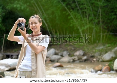 Beautiful and attractive young Asian female taking a picture of the beautiful forest with her retro film camera. Outdoor and camping concept.